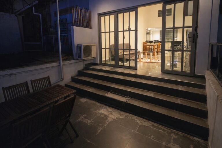 Nighttime view of a private balcony with wooden table and chairs, leading through glass doors into a warmly lit loft-style living and kitchen area.