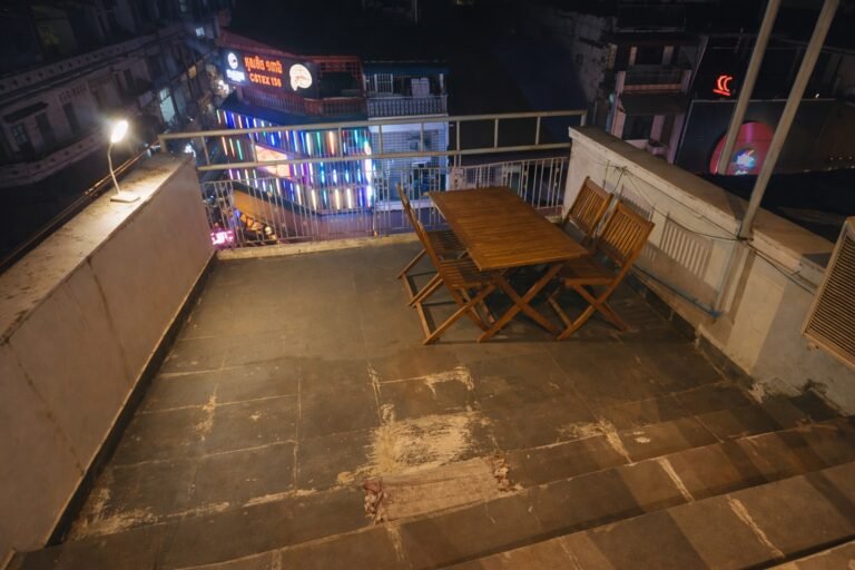 Nighttime view from the apartment balcony showing a wooden outdoor table and chairs, tiled terrace steps, and illuminated city buildings in Phnom Penh below.