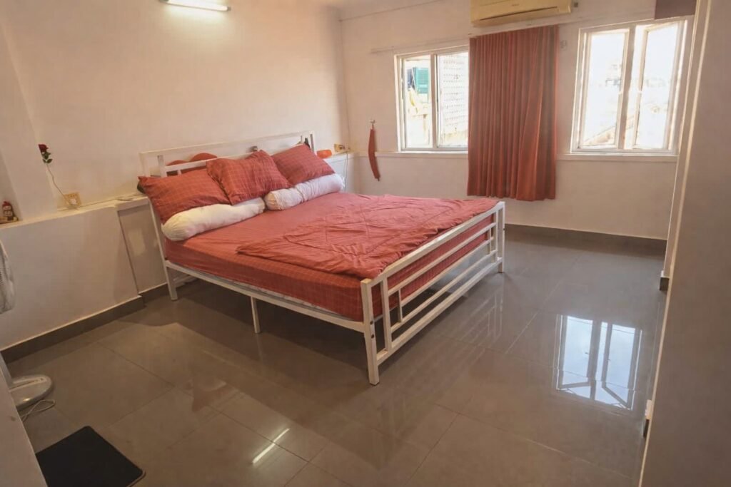 Large bedroom with a white metal bed, red bedding, glossy tiled floors, and two bright windows with curtains.