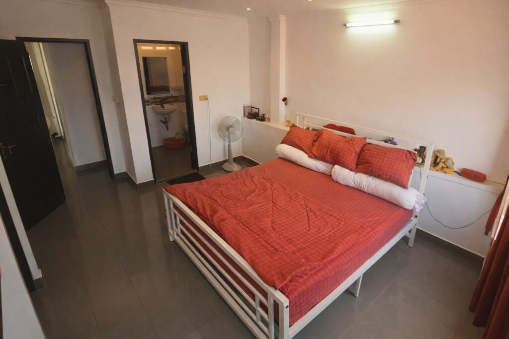Bright bedroom with a white metal bed dressed in red bedding, glossy dark tile floors, and two windows letting in daylight.