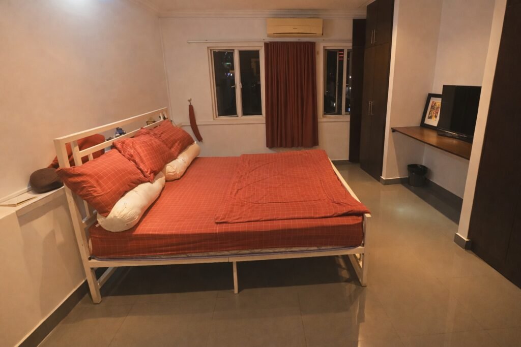 Bright, modern bedroom at night with a red duvet on a white bed frame, tiled floors, built-in dark wardrobes, and a wall-mounted desk with a TV. Caption: Description: